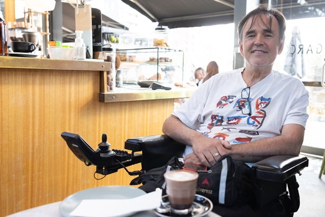 A person who uses a wheelchair sits in a cafe on a sunny day. He has a mocha and is smiling.
