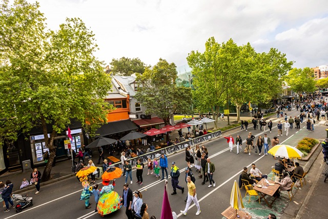 overhead view of a crowd of people walking down the street. the street is lined with green trees and various stalls, some with umbrellas, adding to the festive atmosphere.