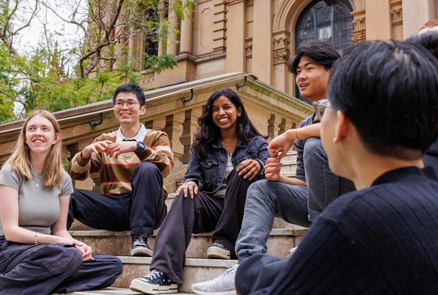 A group of five young adults sit on outdoor stone steps, talking and smiling in front of a historic building.