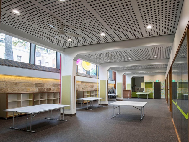 Row of wooden shelves against the wall on the left, two trestle tables standing between vertical columns of illuminated arts centre room. One trestle table in the middle.