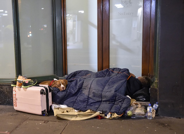 A person sleeping with a dog on the footpath, covered in a sleeping bag and surrounded by a suitcase, clothes and food.
