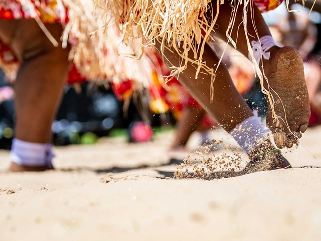Close-up of feet wearing traditional anklets and stepping on sandy ground, with straw skirts visible in the background.