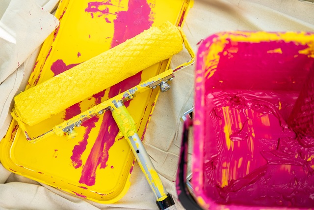A paint roller covered in bright yellow paint rests on a yellow tray next to a bucket of pink paint.