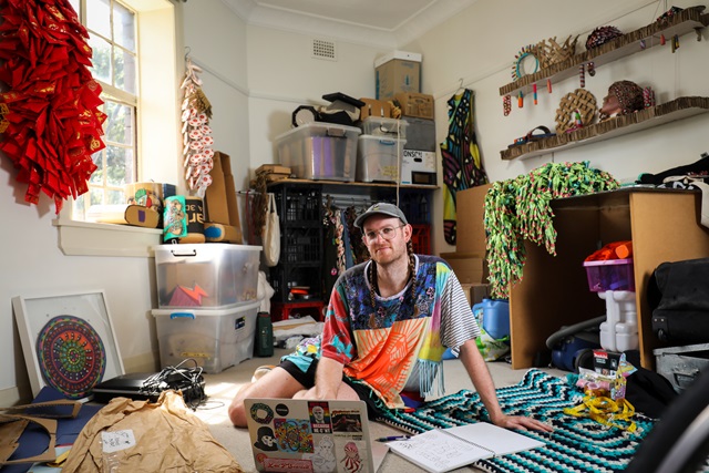 An adult sitting on the floor wearing colourful clothes and surrounded by artwork, a computer and boxes.