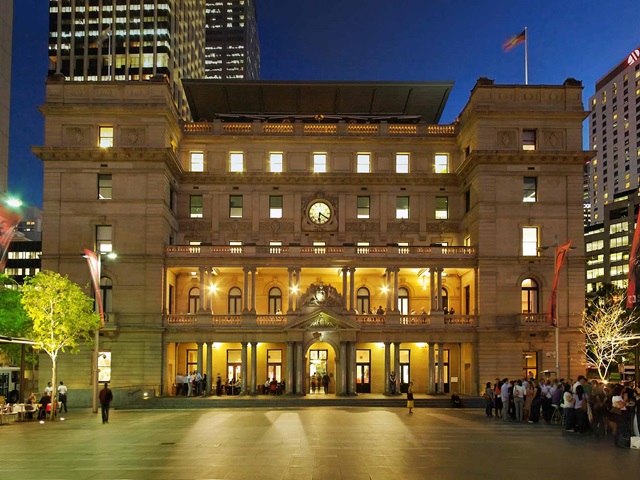 A heritage building with open plaza at twilight. Tall city buildings are in the background. People stand and sit at a cafe bar.