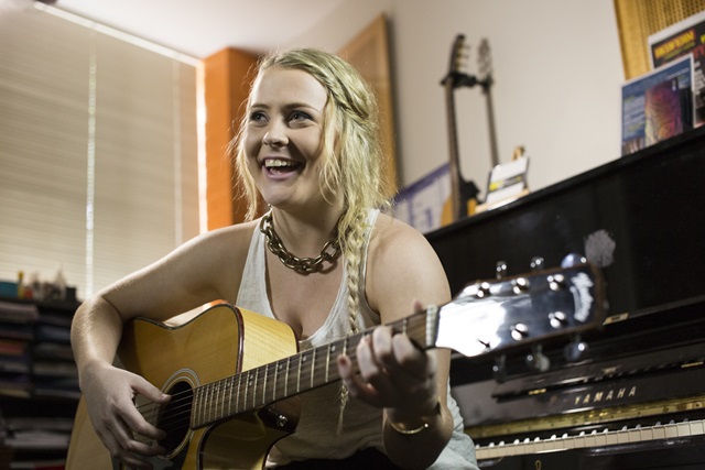 A young woman playing the guitar with a piano behind her.
