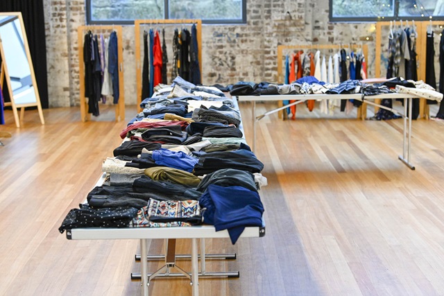 In a large hall are several tables with folded clothes, as well as clothes racks with colourful items.