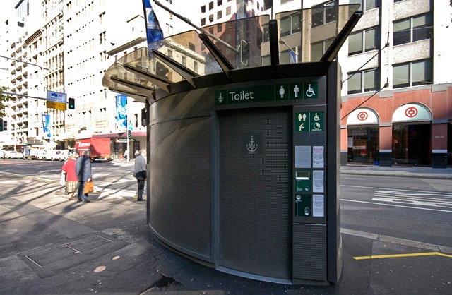 Large automatic public toilet located on a pavement next to a main road