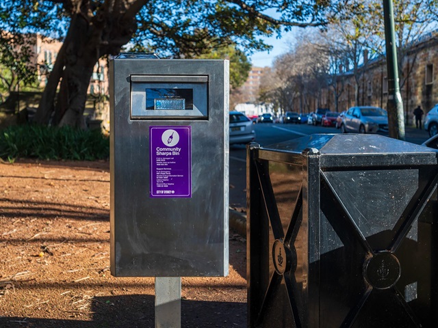A stainless steel community sharps bin placed outdoors. The bin is rectangular and has a small opening at the top for disposing of needles safely, as well as 2 purple stickers on the front of the bin indicating its purpose.