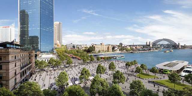 City buildings on a harbour foreshore with a large bridge in the distance. There is an open plaza and trees beside the waterfront.
