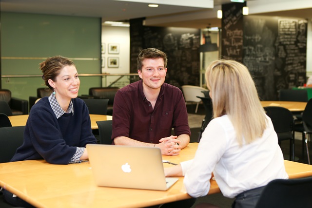Three people at a large table looking at an Apple computer