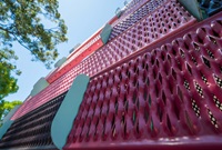 Close-up view of pink perforated metal panels with green brackets, photographed from below, with a tree and blue sky in the background.