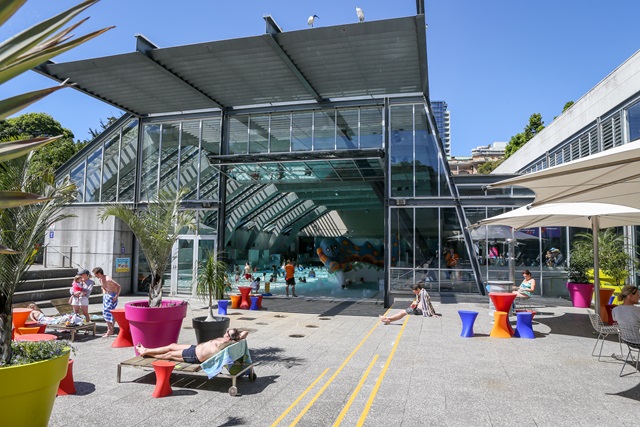People lying on deck chairs, plants in planter boxes and the indoor pool in the distance.