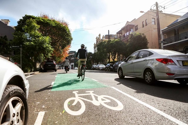 Two cyclists ride on a cycleway between parked cars and a traffic lane.