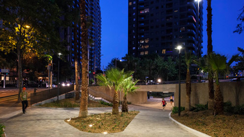 A night time view of the city showing tall buildings in the background and structured paths in the foreground with trees and lighting.