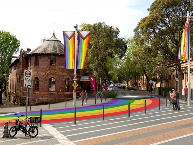 A street features a rainbow-painted crossing with nearby trees and an old brick building. Rainbow flags are displayed on poles.