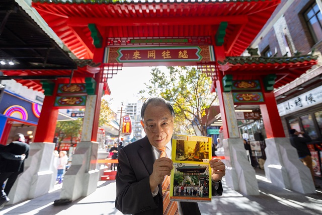 A man in a suit holds up an historical photo in front of a red Chinese-style gate in a busy outdoor area.
