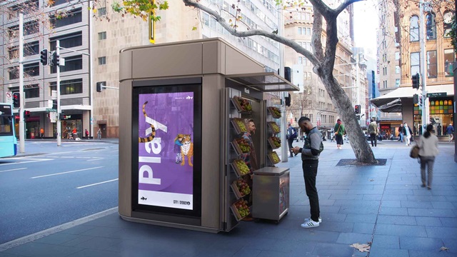 A fruit stand kiosk on a city pavement. A digital screen is on one wall.