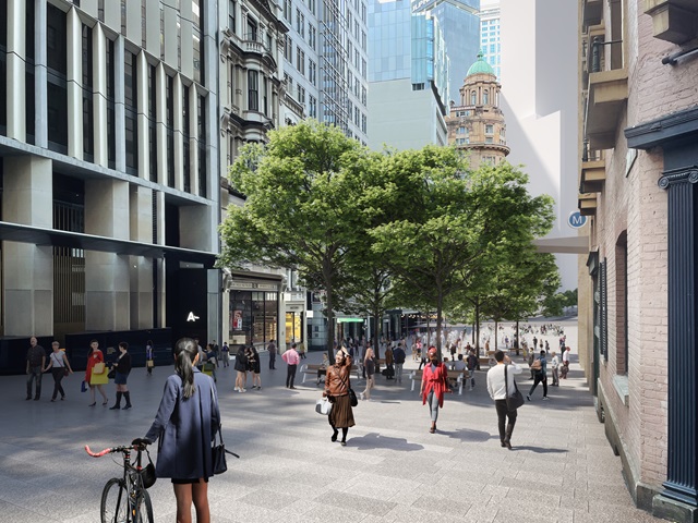 The street is paved for people walking with seats under large trees, surrounded by tall buildings. Many people are walking round through the pedestrianised area.
