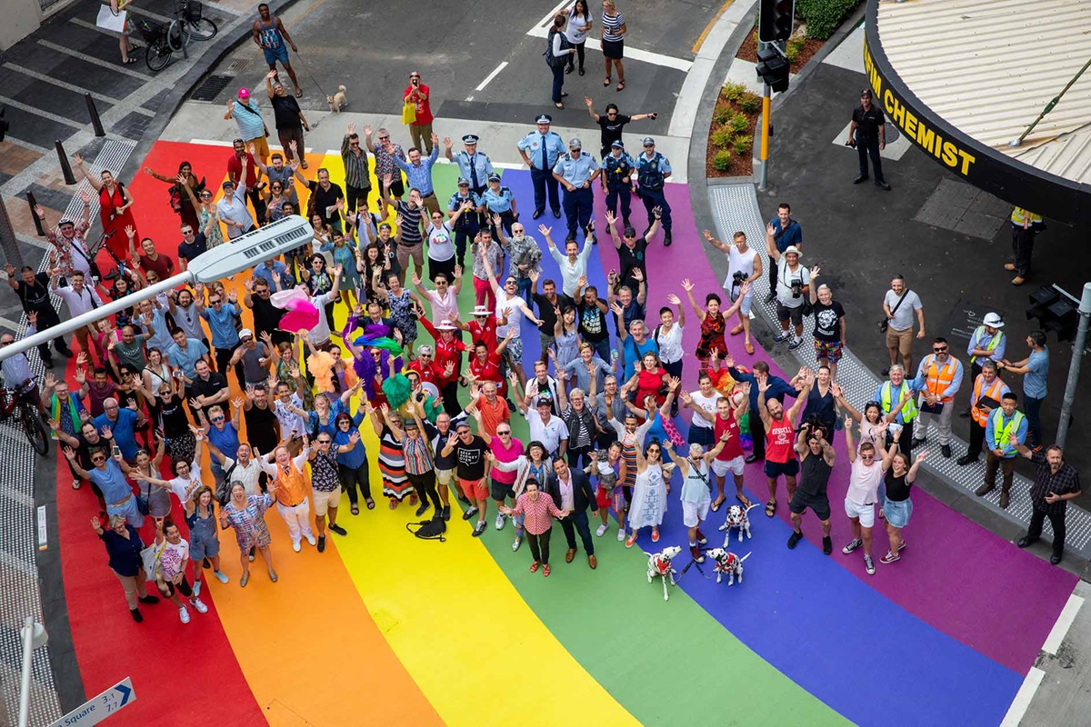 Drone photograph of people standing on a rainbow-coloured street crossing.