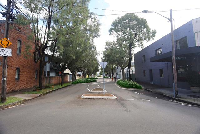 A view down a street with a chicane and foliage verges. There is a 'Give Way' sign on a pedestrian traffic island in the foreground, and two bikers travelling up the street in the distance.