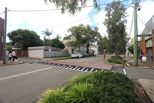 A four-way intersection with a raised traffic threshold at its centre. The streets have foliage verges and footpaths. Several pedestrians and bikers are using the footpaths and streets, respectively.