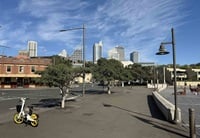 A mostly empty street with trees, a parked yellow hire bike, and city buildings in the background under a blue sky with wispy clouds.