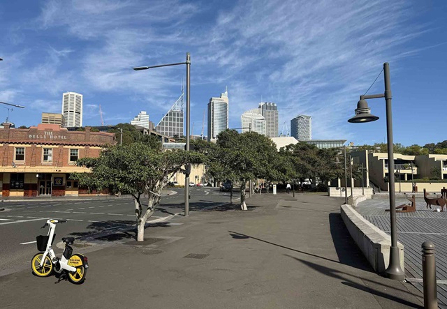 A mostly empty street with trees, a parked yellow hire bike, and city buildings in the background under a blue sky with wispy clouds.