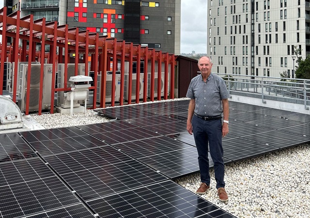 A person stands on a rooftop among several rows of solar panels. Red structure and buildings in the background.