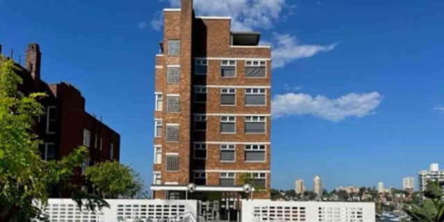 A tall brick apartment building with multiple windows under a blue sky. White decorative barriers in the foreground. Distant cityscape in the background.