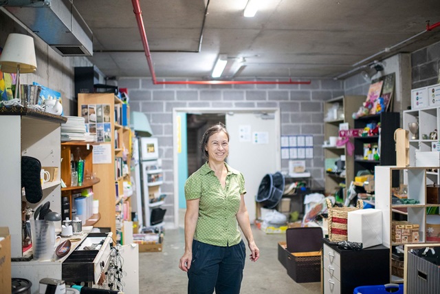 a person stands in a brick wall storage room filled with shelves, boxes, papers and other objects scattered around. Fluorescent lights and a red pipe on the ceiling.