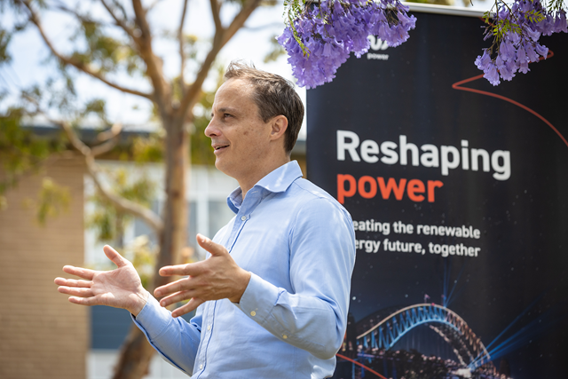 Person wearing professional attire stands in and talks in front of a banner. The banner includes text: reshaping power and creating the renewable energy future, together.