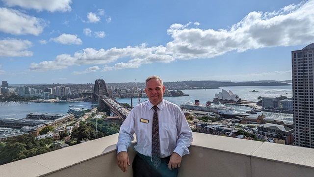 A person stands on a rooftop, leaning against the railing. Sydney harbour and the Sydney Harbour Bridge in the background.