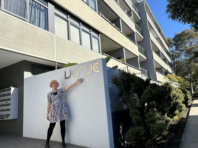 A person with short hair standing outside a modern building. They are touching a large, wall-mounted sign that reads “Verve 20-41.” The building has multiple floors with balconies and visible windows. Small trees and shrubs surround the entrance, and the sky is clear.