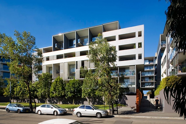 A modern apartment façade with recessed balconies and a lawn area with trees in front. The building is several storeys tall.
