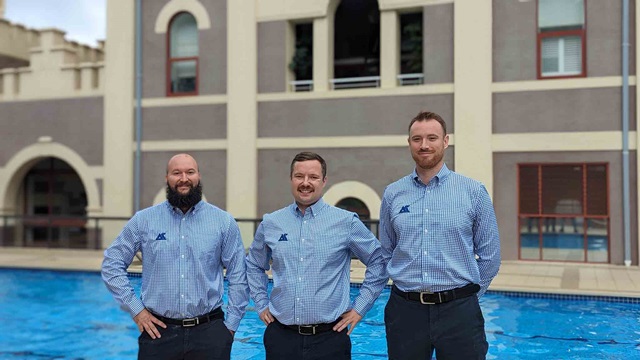 Three people wearing a shirt and pants uniform set. They stand in front of a building with a pool in the foreground.