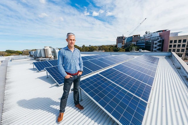 A person stands on a rooftop with solar panels, with city buildings, trees, and a crane visible in the background under a partly cloudy sky.