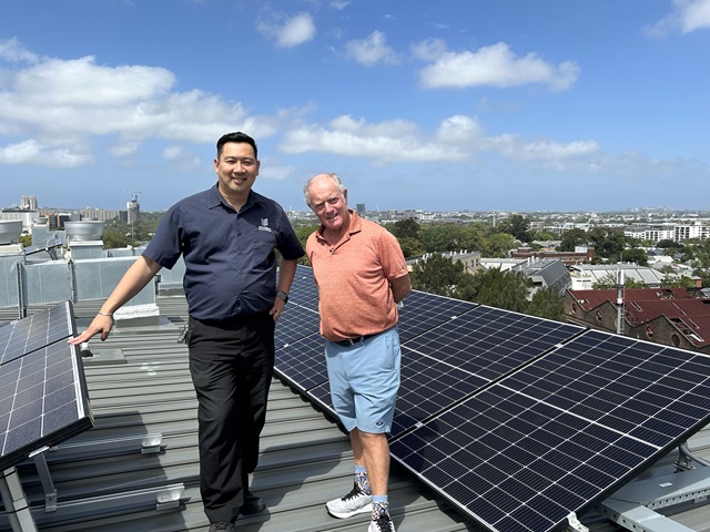 Two men stand on a rooftop next to solar panels, with a cityscape visible in the background under a partly cloudy sky.