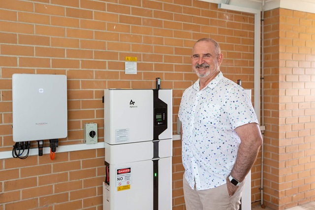A person stands next to a wall-mounted solar inverter and battery storage system. The equipment is installed on a brick wall, with various warning labels and an electrical switchboard.