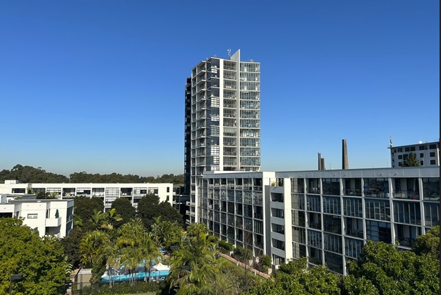 Tall modern apartment building with glass windows, surrounded by smaller buildings and greenery under a clear blue sky.