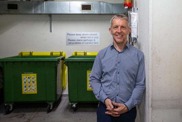 A smiling man in a blue and white checked shirt stands in front of two large recycling bins in an indoor building bin area.