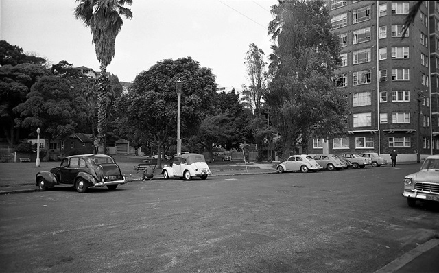 An historic photo of cars parked and trees and tall building.