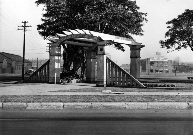 An historic photo of a road, footpath and entrance to a park.