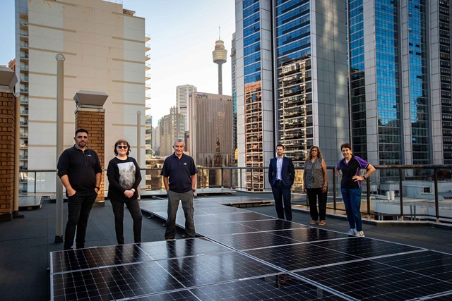 Six men and women stand on a city rooftop that has solar panels.