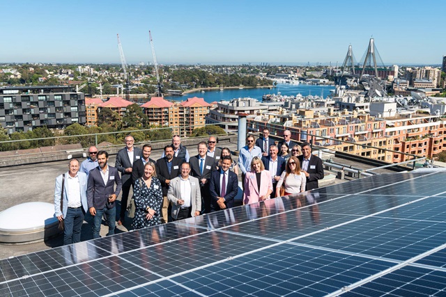 Group of people stand on a city rooftop with solar panels. Anzac Bridge, bay and buildings in the background.