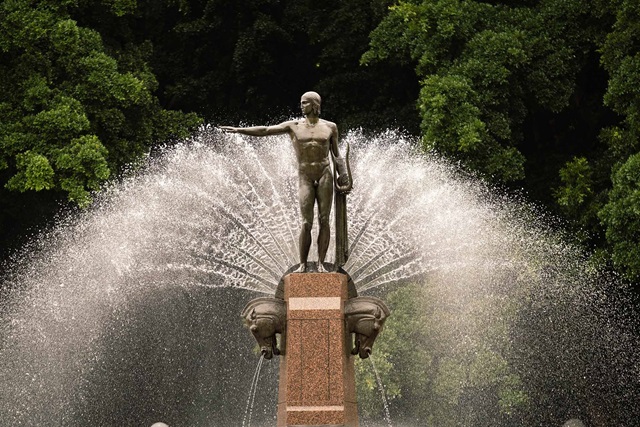 Water jets spaying a fan-like shape around statue on top of the Archibald Memorial Fountain. Trees in background.