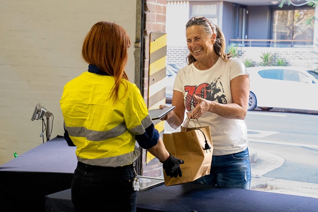 A worker in high-vis is handed a laptop and a full paper carry bag with a power cord sticking out.