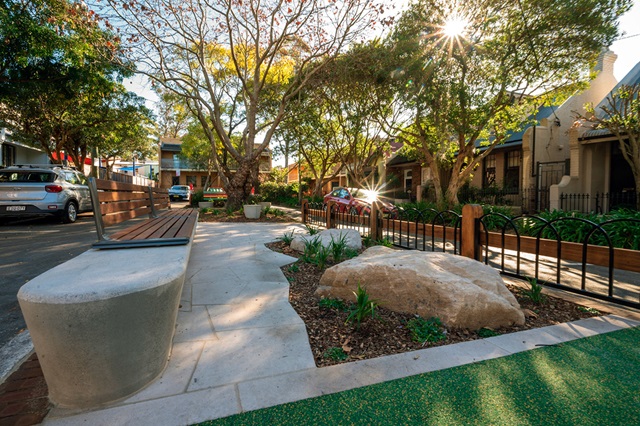 A sunlit urban park with a curved bench, large rock, and trees. A car is parked nearby, and houses are visible in the background.