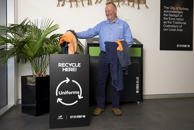 A person stands next to a uniform recycling bin, holding uniforms. A sign on the wall acknowledges the Gadigal of the Eora nation as Traditional Custodians of the area.
