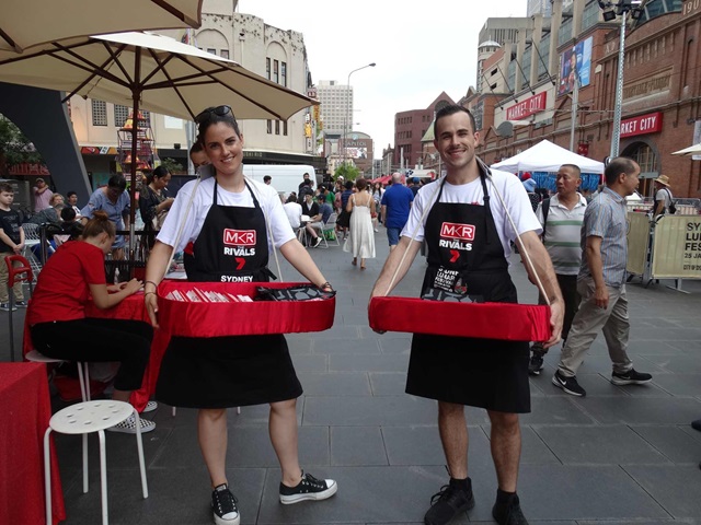 2 adults wearing My Kitchen Rules branded aprons handing out promotional materials outside Market City